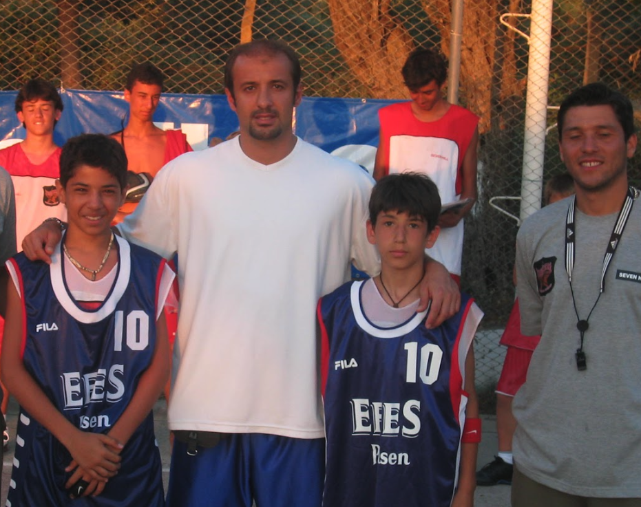 Açık hava basketbol sahasında mavi formalarıyla yetişkinlerle birlikte hatıra fotoğrafı çektiren iki çocuk