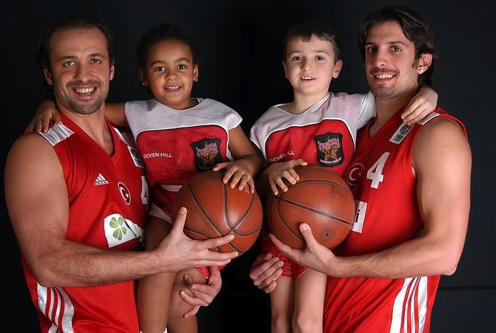 Kırmızı basketbol formalarıyla iki sporcu ve iki çocuk, basketbol toplarıyla birlikte