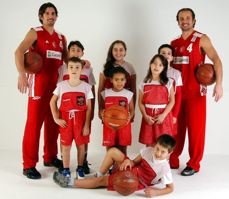 Kırmızı beyaz basketbol formalarıyla iki yetişkin ve çocukların toplarla birlikte poz verdiği grup fotoğrafı