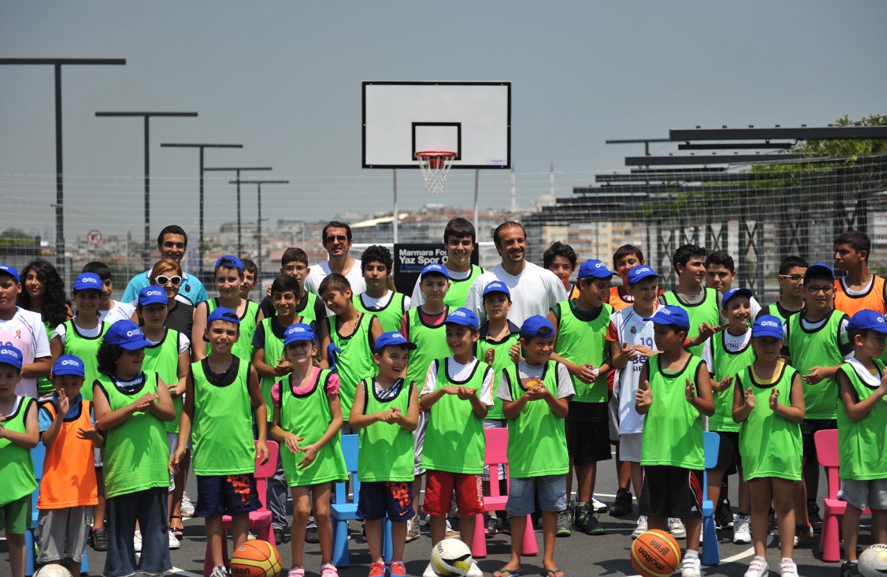 Açık hava basketbol sahasında yeşil antrenman yelekleri ve mavi şapkalarıyla bir araya gelen çocuklar ve yetişkinler